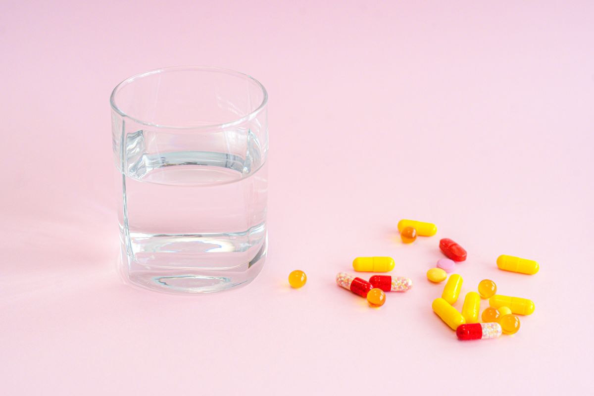 A glass of water beside an assortment of colorful pills on a pink background.
